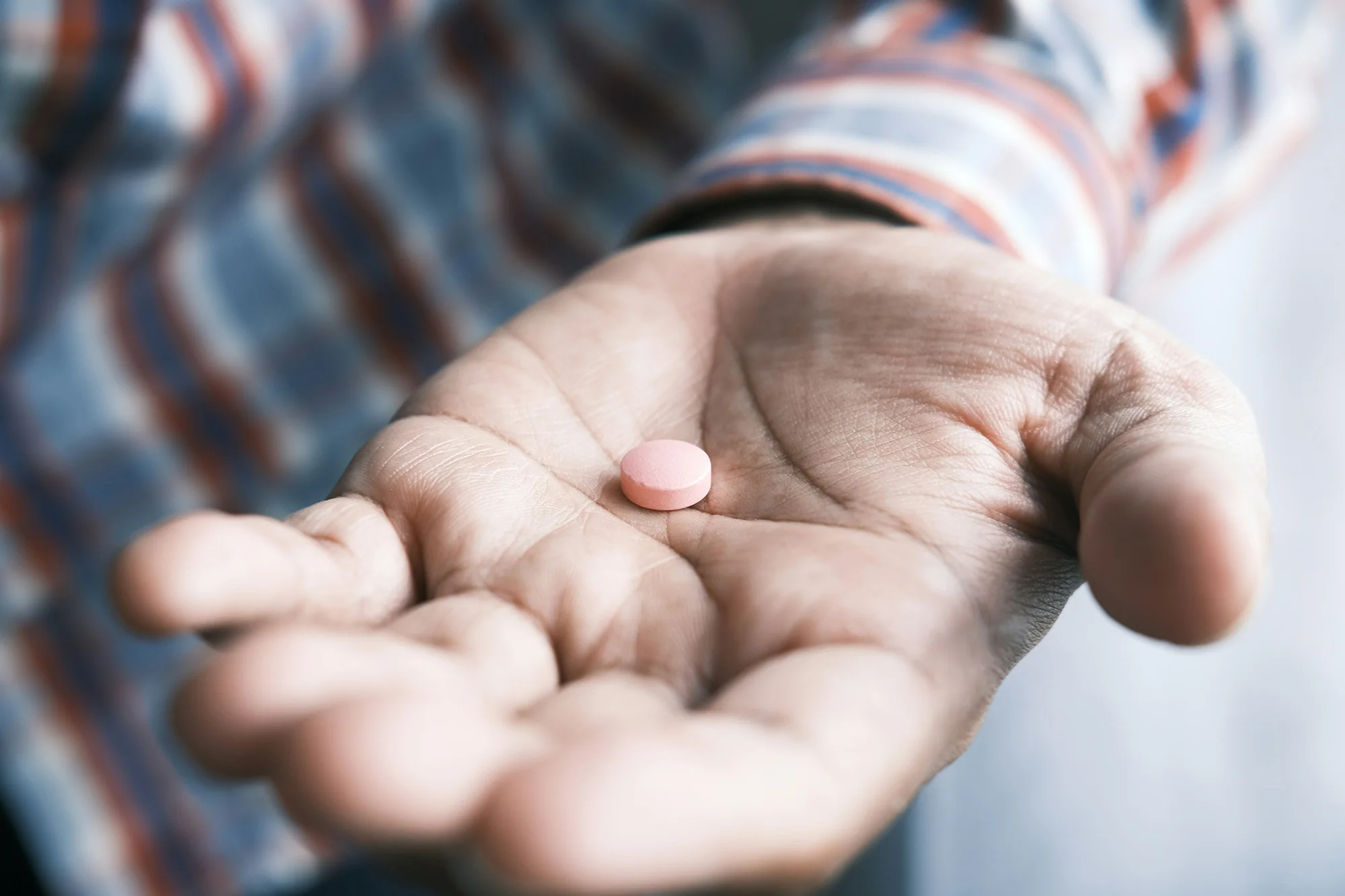 closeup photo of the hand of a man wearing a plaid shirt, holding a single orange pill in his palm